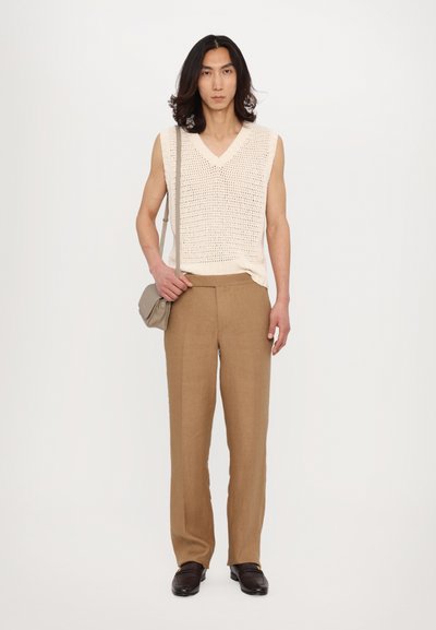 Gilet en maille beige clair avec un col en V, associé à un pantalon tailleur marron clair. La tenue est complétée par des mocassins marron foncé et un petit sac.