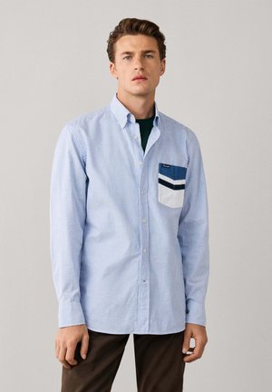 Joven con el pelo corto y castaño que lleva una camisa de rayas azules y blancas con botones y un bolsillo multicolor en el pecho, de pie frente a un fondo liso.