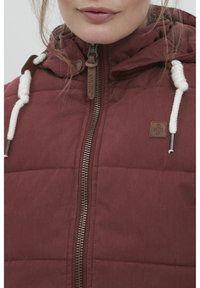 Veste isolée bordeaux avec fermeture éclair sur le devant, capuche et cordons blancs. Présente un patch de la marque et une texture matelassée.