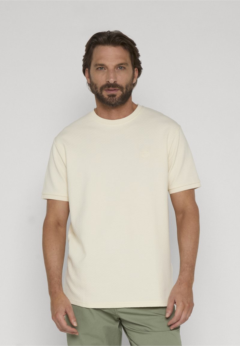 Homme barbu portant un t-shirt texturé crème uni à manches courtes et un pantalon vert olive, debout devant un fond gris clair.