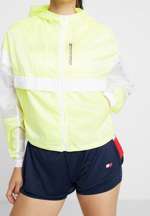 Femme portant une veste à capuche jaune fluo zippée et un short de sport en mesh bleu marine avec des détails rouges et blancs.