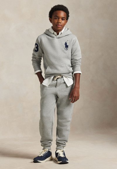 Garçon debout, mains dans les poches, portant un sweat à capuche gris, un pantalon de jogging assorti, une chemise blanche et des baskets bleues sur un fond uni.