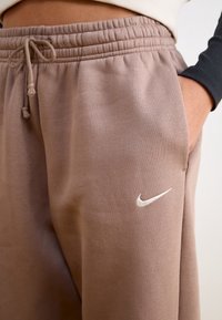 Close-up of taupe sweatpants with elastic waistband, drawstring, side pocket, and white Nike swoosh logo on thigh.