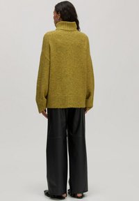 Geelgroene oversized coltrui met geribbelde zoom, gedragen met wijde zwarte leren broek en franjeschoenen in het zwart.