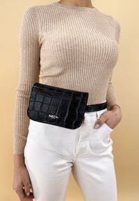 Czarna torba z ekoskóry w wzór krokodyla z klapą, noszona w talii. Beżowa, prążkowana bluzka z długim rękawem i białe dżinsy.