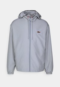 Chaqueta con capucha y cremallera en gris claro, fabricada en tejido suave, con puños elásticos y un pequeño logo bordado en el lado izquierdo del pecho.