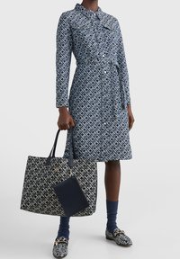 Kobieta w niebieskiej sukience z wzorem z guzikami oraz dopasowaną torbą na ramię w tym samym wzorze i mokasynami, stojąca na tle jednolitym.