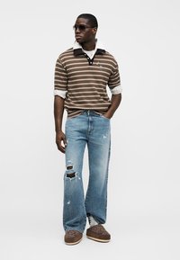 Καφέ ριγωτό πόλο πουκάμισο με γκρι λεπτομέρειες, φθαρμένο ανοιχτό μπλε τζιν και καφέ παντόφλες. Το μοντέλο φοράει γυαλιά ηλίου και στέκεται με το ένα χέρι στη μέση.