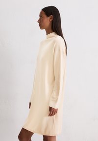 Gebreid beige jurk met een hoge kraag, lange mouwen en een relaxte pasvorm, met een zachte textuur en halverwege de bovenbenen.