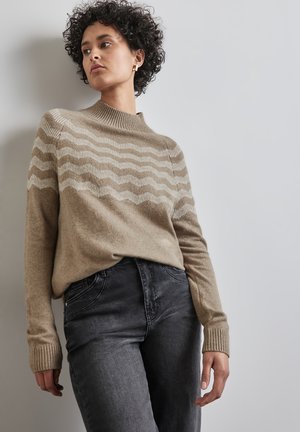 Lyst brun striktrøje med beige bølgemønster, der har høj hals og ribbede manchetter, parret med mørkegrå tætsiddende jeans.