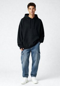 Zwarte hoodie met capuchon en een buidelzak aan de voorkant, gedragen met lichtblauwe cargobroek. Model staat in witte sneakers tegen een effen achtergrond.
