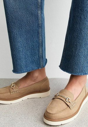 Mocassins en daim marron clair avec un accent métallique décoratif, semelle crème et surface texturée. Portés avec un jean en denim bleu coupe courte.