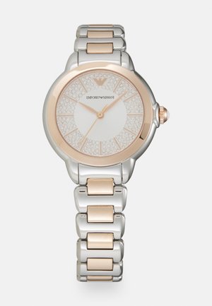 Srebrny i różowozłoty zegarek metalowy z okrągłą tarczą ozdobioną kryształami. Posiada białą tarczę z indeksami godzinowymi i świecącą koronką.