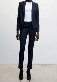 Marineblauw getailleerd colbert en bijpassende slim-fit broek, gecombineerd met een witte top met ronde hals en zwarte puntige enkellaarzen. Soepele stoftextuur.