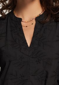 Femme portant un haut noir brodé avec un décolleté en V et deux colliers superposés, l'un en perles multicolores et l'autre en chaîne dorée avec de petites perles.