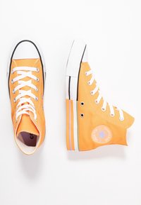Deux baskets montantes Converse orange avec des lacets blancs et des semelles en caoutchouc, l'une vue de dessus, l'autre de côté sur fond blanc.