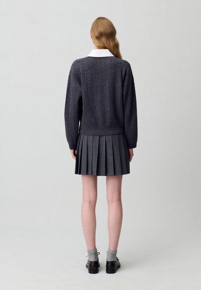 Maglione grigio a trecce con colletto bianco abbinato a una gonna grigia plissettata, calzini grigi e scarpe nere, visto da dietro.