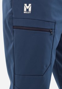 Pantalon de plein air bleu marine en tissu doux ; doté d'une poche zippée et d'une texture lisse ; marqué d'un logo "Millet" blanc.