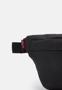 Mustekka kankainen pussi suorakaiteen muotoinen ja teksturoitu pinta. Varustettu punaisella kankaisella tarralla ja mustalla hihnalla sivussa.