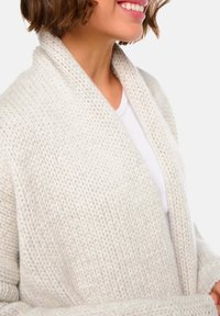 Large cardigan en maille grise clair avec un col châle, présentant un motif texturé et des manches amples, porté sur un haut blanc.