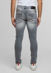 Férfi slim-fit, megfakult szürke farmernadrágban, fehér sportcipőben, narancssárga sarokbetétekkel, egy sima világosszürke háttér előtt áll.