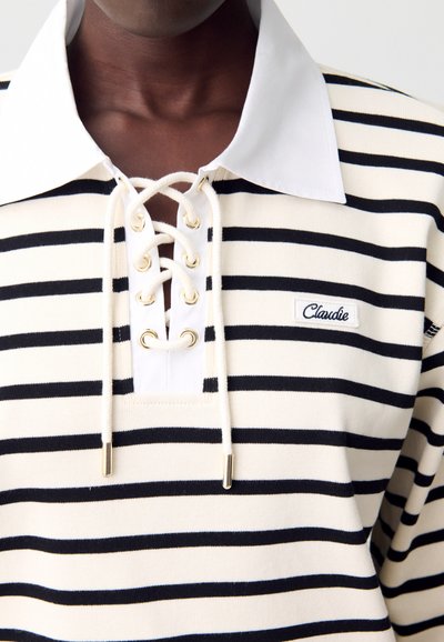 Polo a righe in crema e nero. Presenta un colletto bianco, chiusura con lacci e occhielli dorati, e logo "Claudie" ricamato.