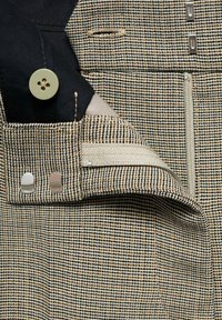 Tissu texturé en taupe et noir, avec un bouton vert, une quincaillerie argentée et une poche zippée avec une doublure contrastée.