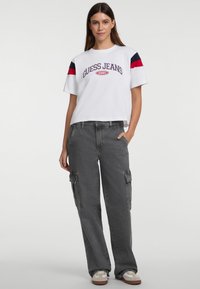 Biely skrátený tričko s červenými a námorníckymi pruhmi na rukávoch a grafikou "GUESS JEANS 1981". Voľné šedé cargo nohavice s bočnými vreckami.
