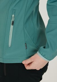 Teal pehmeäkuorijakku, jossa on teksturoitu pinta, puoleen vetoketjuun suljettava tasku, elastiset hihansuut ja logotunnus helman lähellä. Ominaisuuksiin kuuluu muotoiltu istuvuus.
