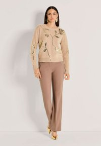 Frau in einem beigen Pullover mit goldenen Blumenmuster, taupefarbener, weit geschnittener Hose und goldenen spitzen Pumps, vor einem schlichten Hintergrund stehend.