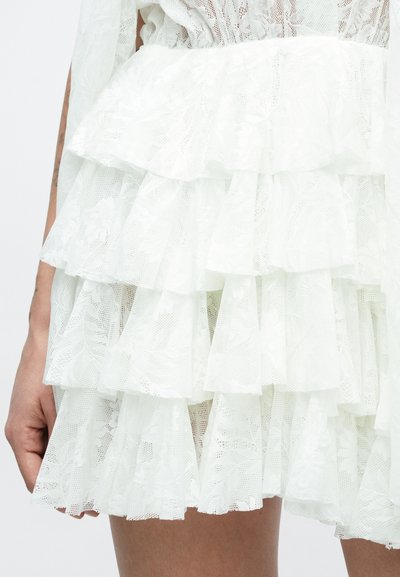 Biela vrstvená čipkovaná sukňa s kvetinovými vzormi, ktorú nosí osoba, vidieť ruku a hornú časť stehna na jednoliatej pozadí.