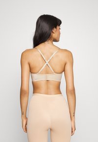 Beige bh med en sømløs design, justerbare stropper som krysser bak og krokfeste. Glatt tekstur med minimale hardware detaljer.