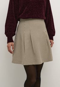 Jupe plissée à carreaux beige, fabriquée en tissu texturé. Portée avec un pull bourgogne ajusté et des collants noirs.