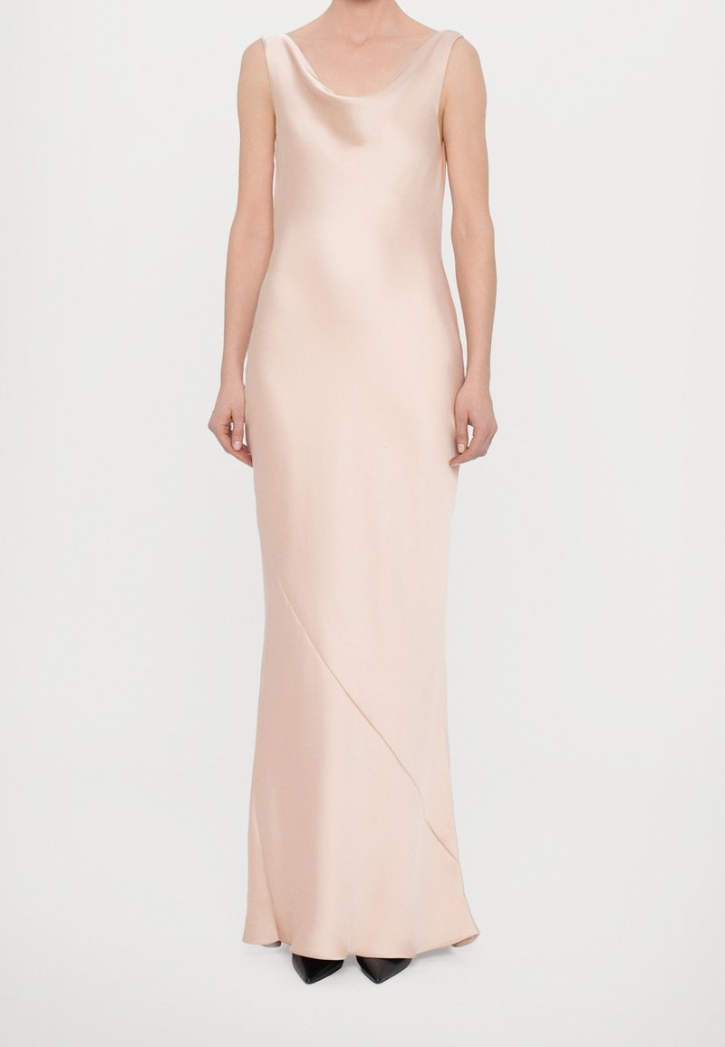 Femme portant une robe en satin rose clair sans manches avec un décolleté drapé et des chaussures noires pointues sur un fond uni.