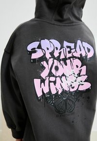 Un hoodie noir avec un grand graphique coloré portant l'inscription "FAIS VOLER TES AILES" en rose et blanc, avec un papillon en dessous.