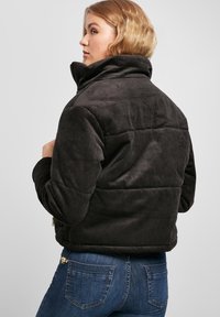 Zwarte corduroy pufferjack met een hoge kraag, geribbelde textuur en ritssluiting, gedragen over blauwe jeans met een metallic accent.