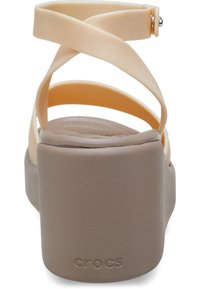 Sandales Crocs beige clair avec des brides croisées, une semelle rembourrée et un talon plateforme marron uni. Logo visible sur le talon.