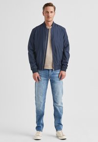 Marineblaue Bomberjacke mit glatter Textur, gerippten Bündchen und Reißverschluss, kombiniert mit hellblauen Jeans und beigen Sneakers.