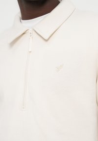 Κοντινό πλάνο ενός ατόμου που φοράει κρεμ πουκάμισο με φερμουάρ και γιακά, με ένα μικρό κεντημένο λογότυπο πουλιού στο στήθος.