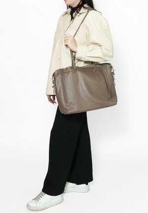 Borsa in pelle marrone con doppi manici, tasche laterali con zip e finitura testurizzata, indossata da una persona con una camicia chiara e pantaloni neri.