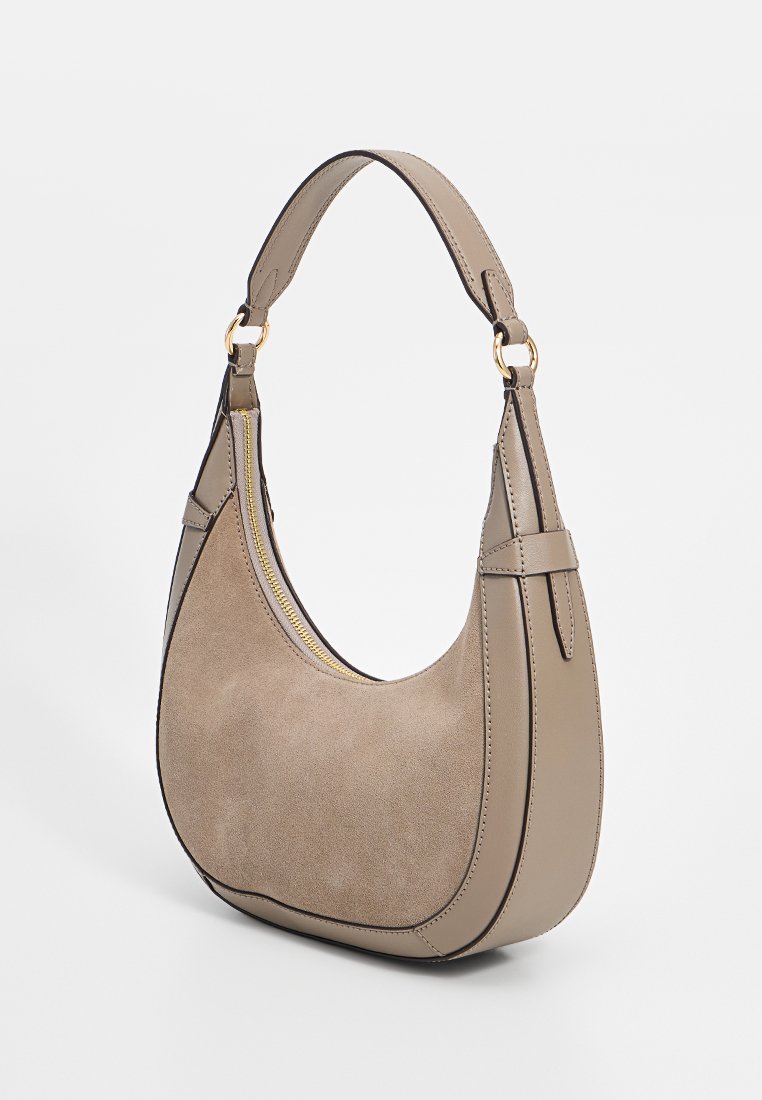 Beige suède handtas met een gebogen ontwerp, gouden hardware en een verstelbare schouderriem. Bevat een ritsluiting en contrasterende stikdetails.