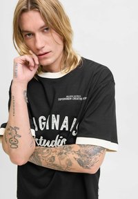 Camiseta negra de manga corta con letras en blanco y puños acanalados de color crema. Presenta tatuajes en los brazos y un primer plano de la cara.