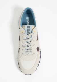 Zapatillas de ante de color claro con acentos azules, cordones texturizados, bordes con ondas y una suela de goma oscura. Marca visible en la lengüeta.