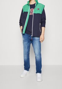 Navy and green zip-up jacket with a high collar, paired with a navy graphic t-shirt and light blue jeans. White sneakers complete the look.