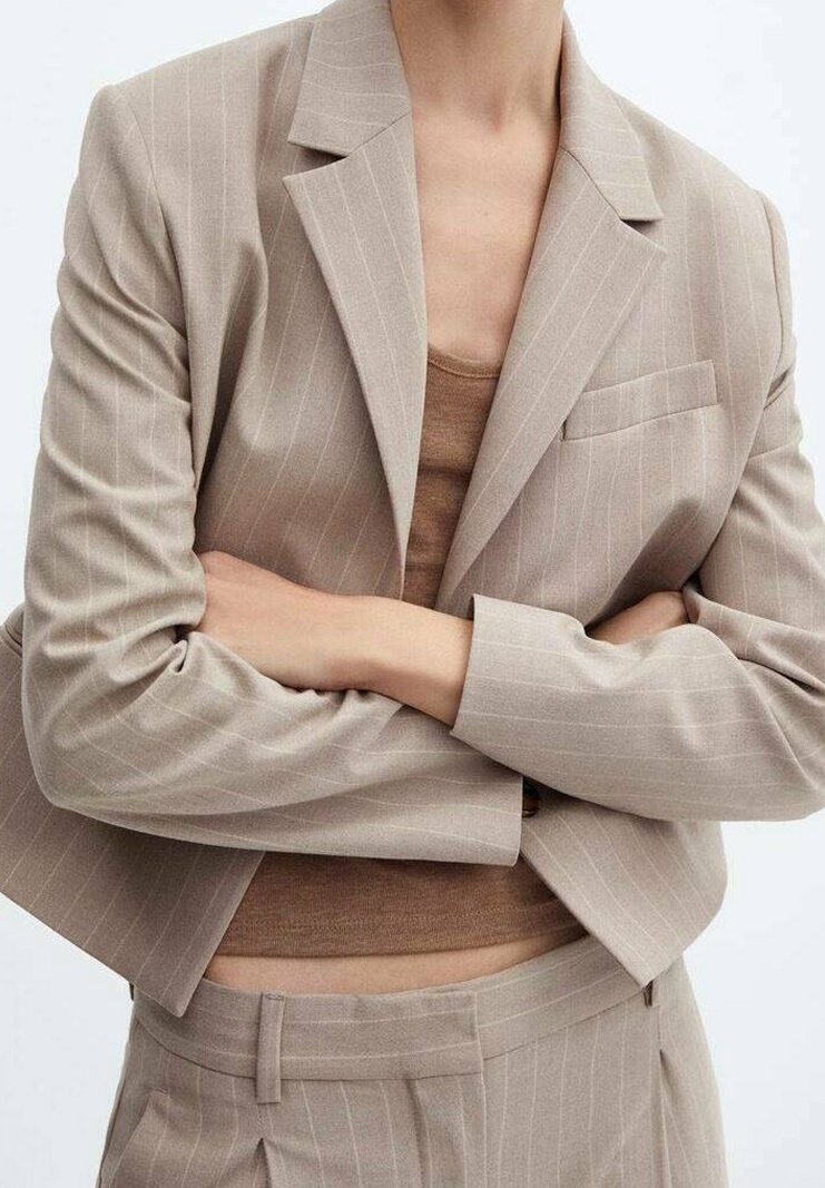 Blazer beige à rayures fines avec une coupe ajustée. Une poche poitrine unique, épaules structurées et manches légèrement raccourcies. Porté avec un haut marron.