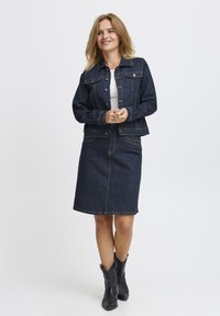 Denim jack en rok set in donkerblauw, met voorzakken, knopen en contrasterende stiksels. Zwarte laarzen maken de outfit compleet.