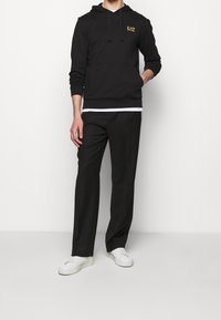 Svart huvtröja med ficka fram och dragsko, kombinerad med löst sittande svarta byxor och vita sneakers. EA7-logotyp i guld.