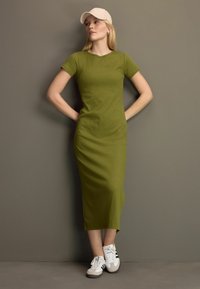 Olijfgroene midi-jurk met korte mouwen en een ronde halslijn, gecombineerd met witte sneakers met zwarte strepen en een beige pet.