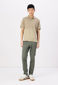 Polo khaki avec un col classique, associé à un pantalon olive ajusté. Le modèle porte des baskets blanches avec des accents beiges. Textures lisses.