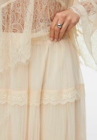 Haut en dentelle crème claire sur une jupe transparente plissée avec bordure en dentelle. Une bague en argent orne une main près de la taille. Textures et motifs délicats.